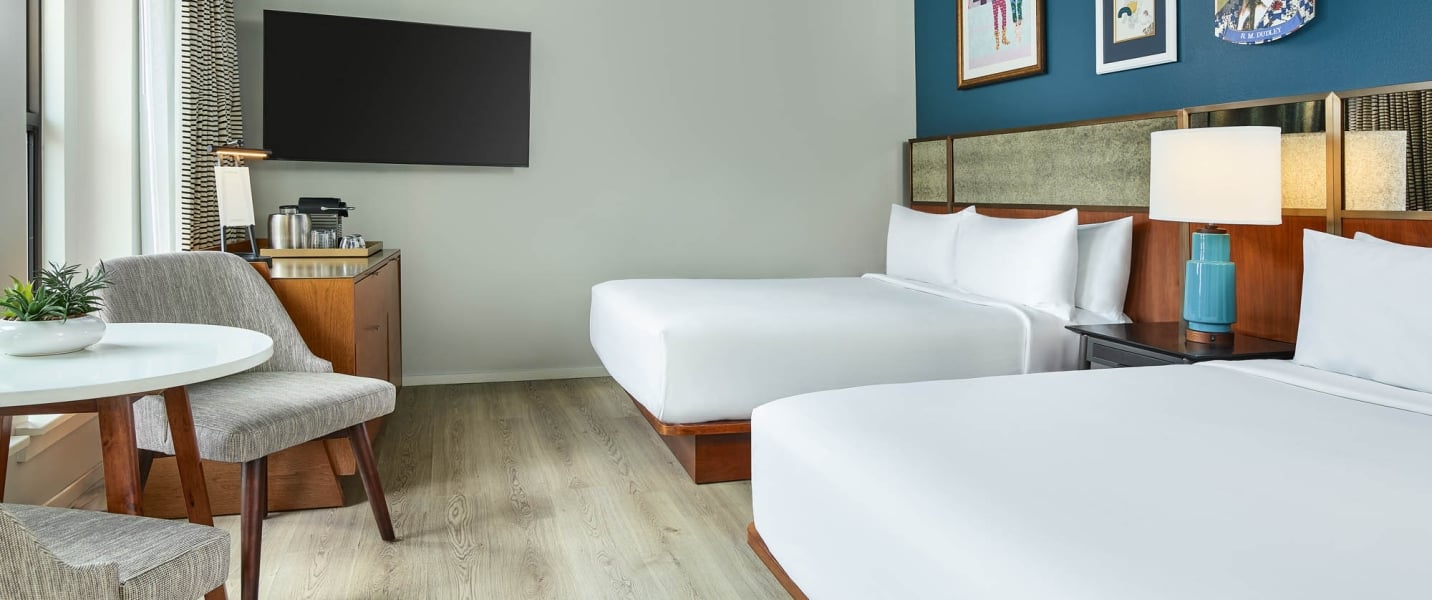 Modern hotel room features two queen beds with white linens, a TV, coffee station, and a round table with a plant and chair, all ready for guests.