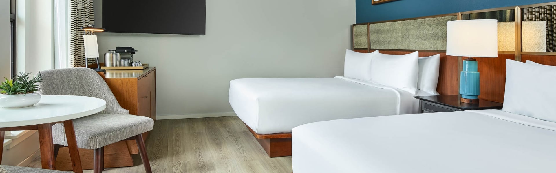 Modern hotel room features two queen beds with white linens, a TV, coffee station, and a round table with a plant and chair, all ready for guests.