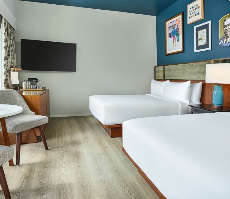 Modern hotel room features two queen beds with white linens, a TV, coffee station, and a round table with a plant and chair, all ready for guests.