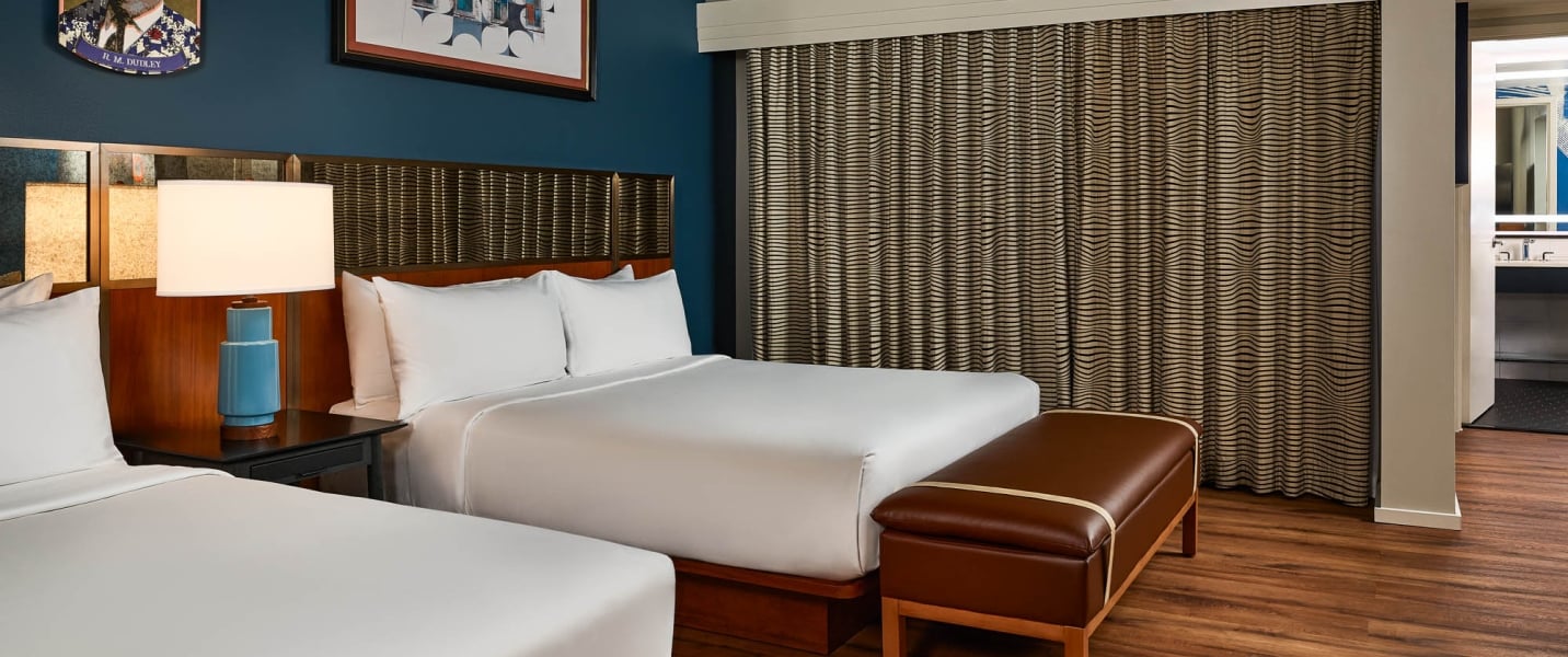 Two queen beds are neatly made with white linens. A brown leather bench, dark blue walls, wood floors, and patterned curtains furnish this modern hotel room.