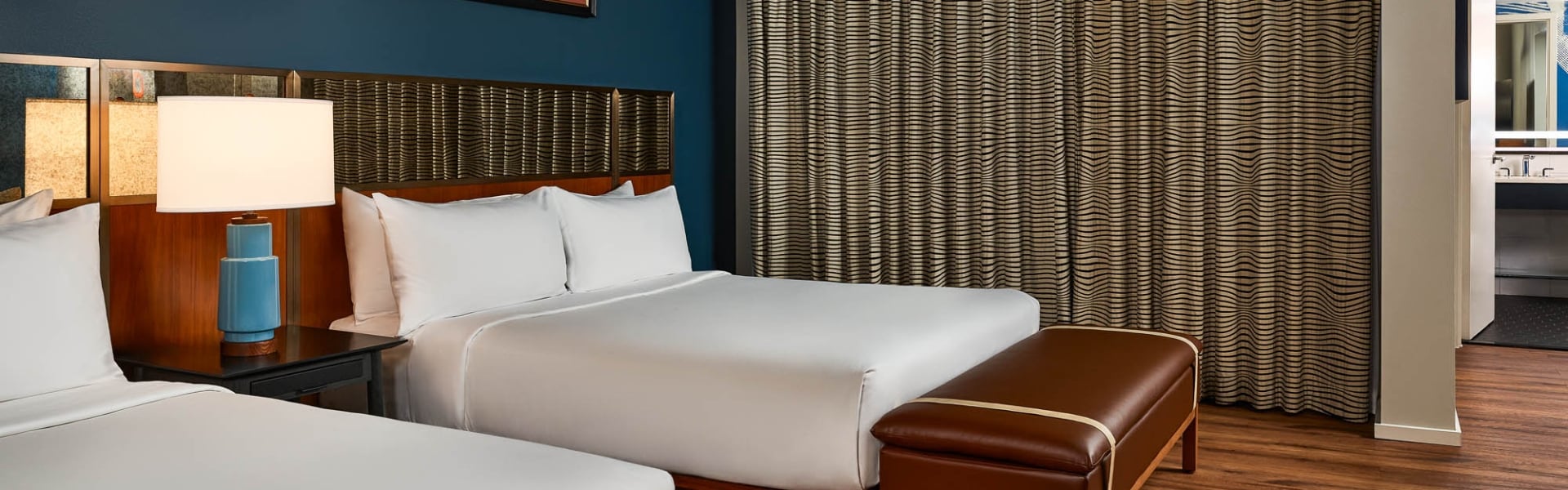 Two queen beds are neatly made with white linens. A brown leather bench, dark blue walls, wood floors, and patterned curtains furnish this modern hotel room.