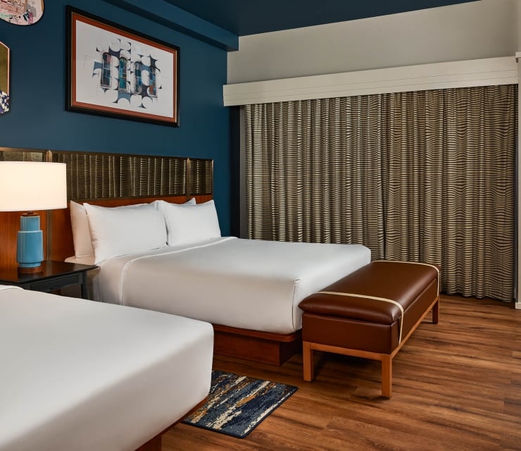 Two queen beds are neatly made with white linens. A brown leather bench, dark blue walls, wood floors, and patterned curtains furnish this modern hotel room.