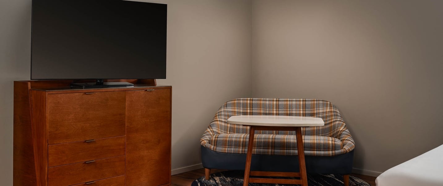 A flat-screen TV rests on a wooden dresser next to a plaid armchair and small table, forming a cozy seating area in an indoor room with light brown walls.