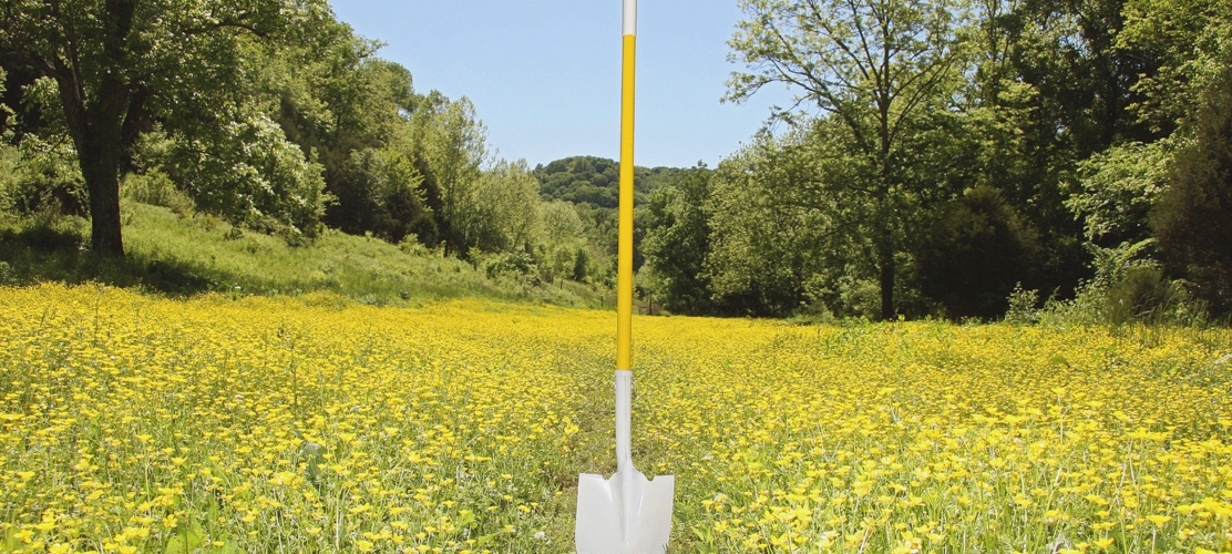 Tall yellow-and-white survey marker standing in a field of yellow flowers with trees in the background.