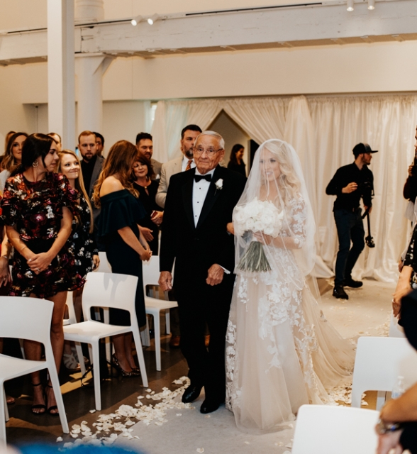 Bride in a lace wedding gown and veil holding a bouquet walks down the aisle with an elderly man in a black tuxedo as guests stand along white chairs. Petals scatter on the floor.