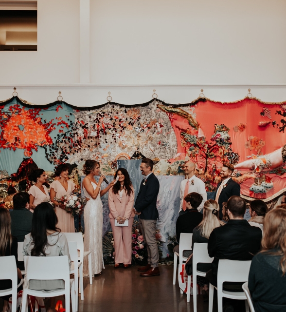 Wedding ceremony with the couple and bridal party standing in front of a large, colorful textile backdrop while guests watch.