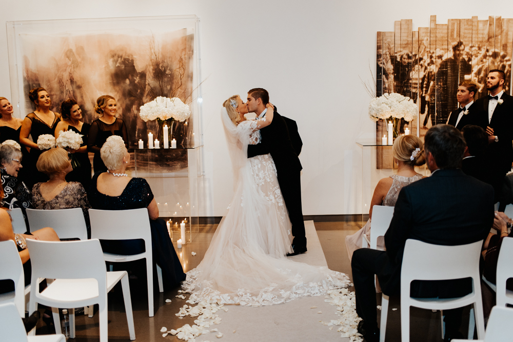 Bride and groom kiss at the altar during a wedding ceremony, with guests seated on white chairs and bridesmaids on the left side.