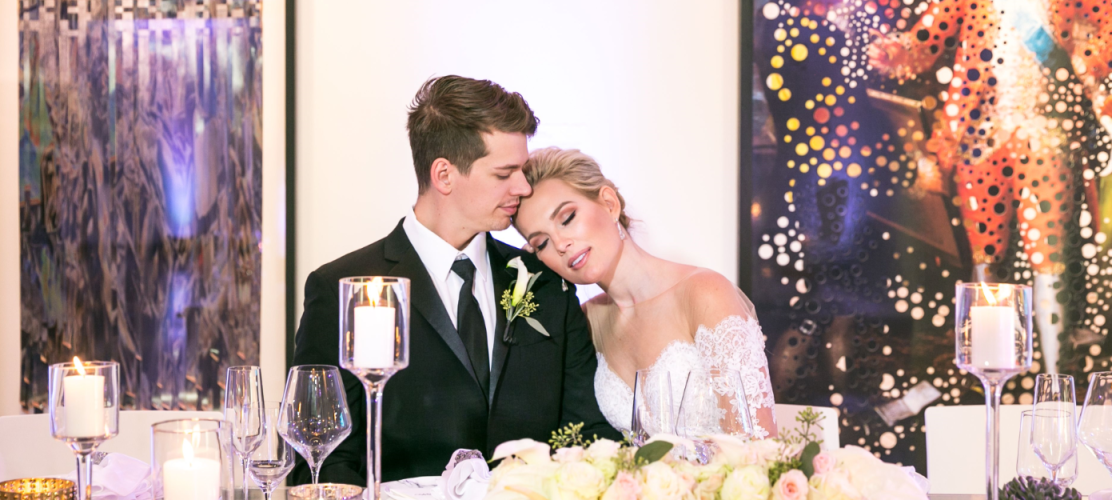 Bride and groom lean their heads together at a candlelit wedding table with white roses and glassware in the foreground