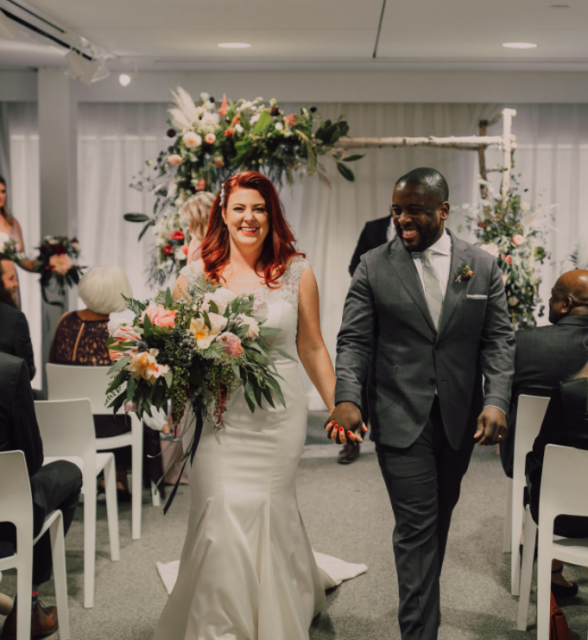 Bride and groom walk down the aisle hand in hand during a wedding ceremony, with guests seated and a floral arch behind them.