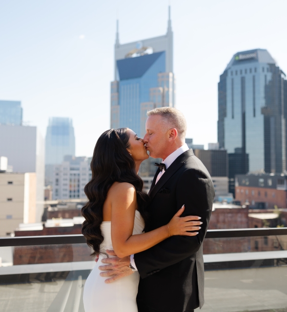 Couple in formal attire kissing on a rooftop with a city skyline behind them.