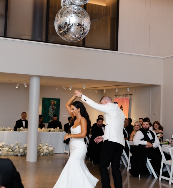 Bride in a white wedding gown and groom in a white suit dance together at a reception as guests look on, with hanging disco balls overhead and a long white-clothed head table in the background.