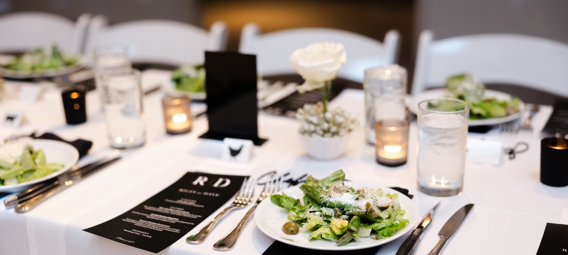 Table set for dinner with salad plates, cutlery, candles, and a white rose centerpiece on a white-tablecloth surface.