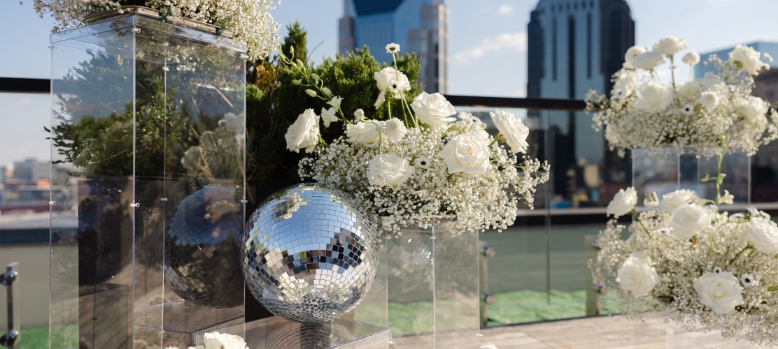 Floral arrangement of white roses and baby's breath with mirrored disco balls on a rooftop railing against a city skyline.