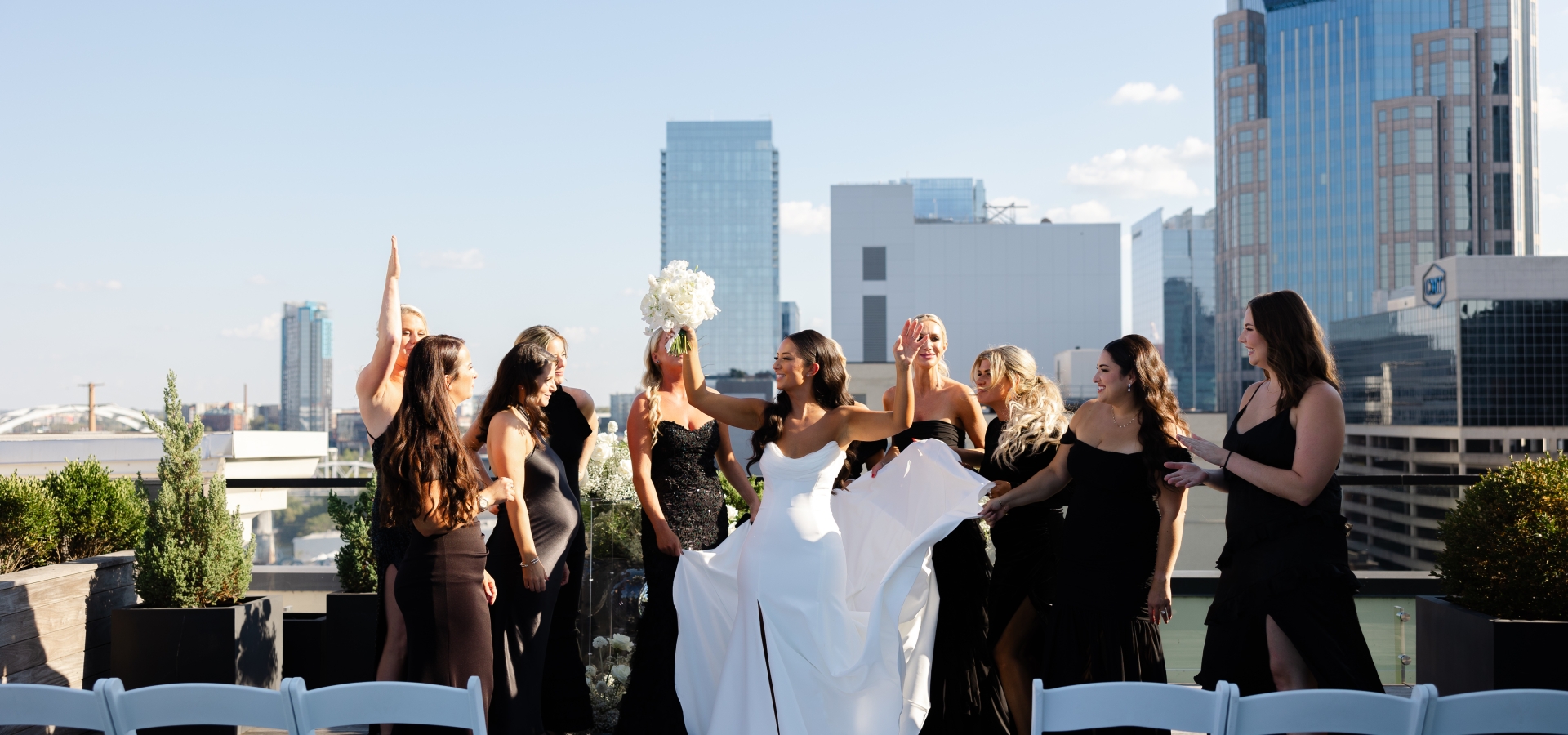 Bride in a white wedding dress lifts her bouquet while surrounded by bridesmaids in black dresses on a rooftop with a city skyline in the background under a clear blue sky.