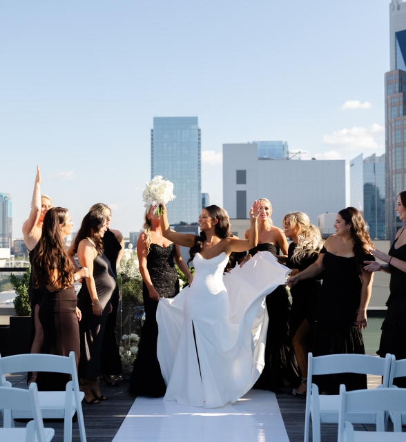 Bride in a white wedding dress lifts her bouquet while surrounded by bridesmaids in black dresses on a rooftop with a city skyline in the background under a clear blue sky.