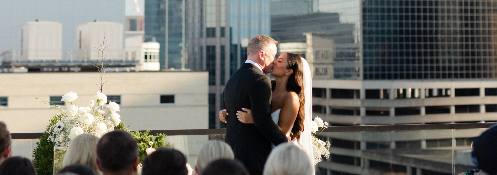 Bride and groom kissing on a rooftop during an outdoor wedding, with guests seated in front and a modern city skyline behind them.